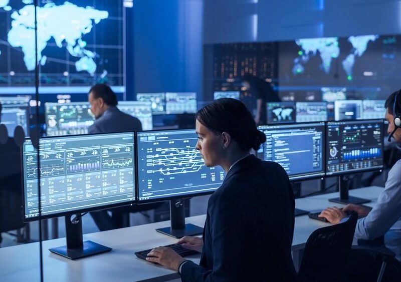 Control room with operators monitoring multiple screens displaying charts and world maps at their desks, blue-toned lighting.