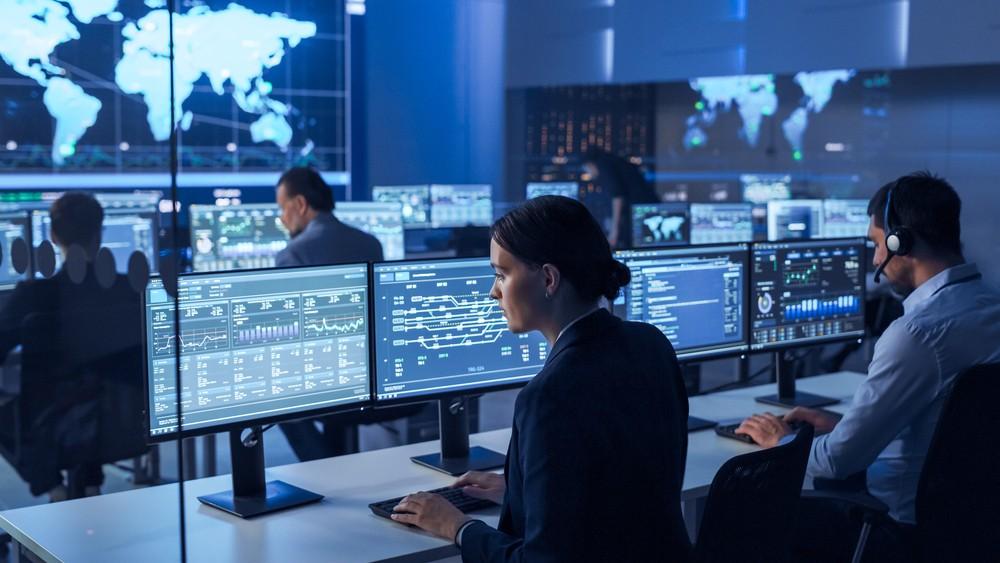 Control room with operators monitoring multiple screens displaying charts and world maps at their desks, blue-toned lighting.